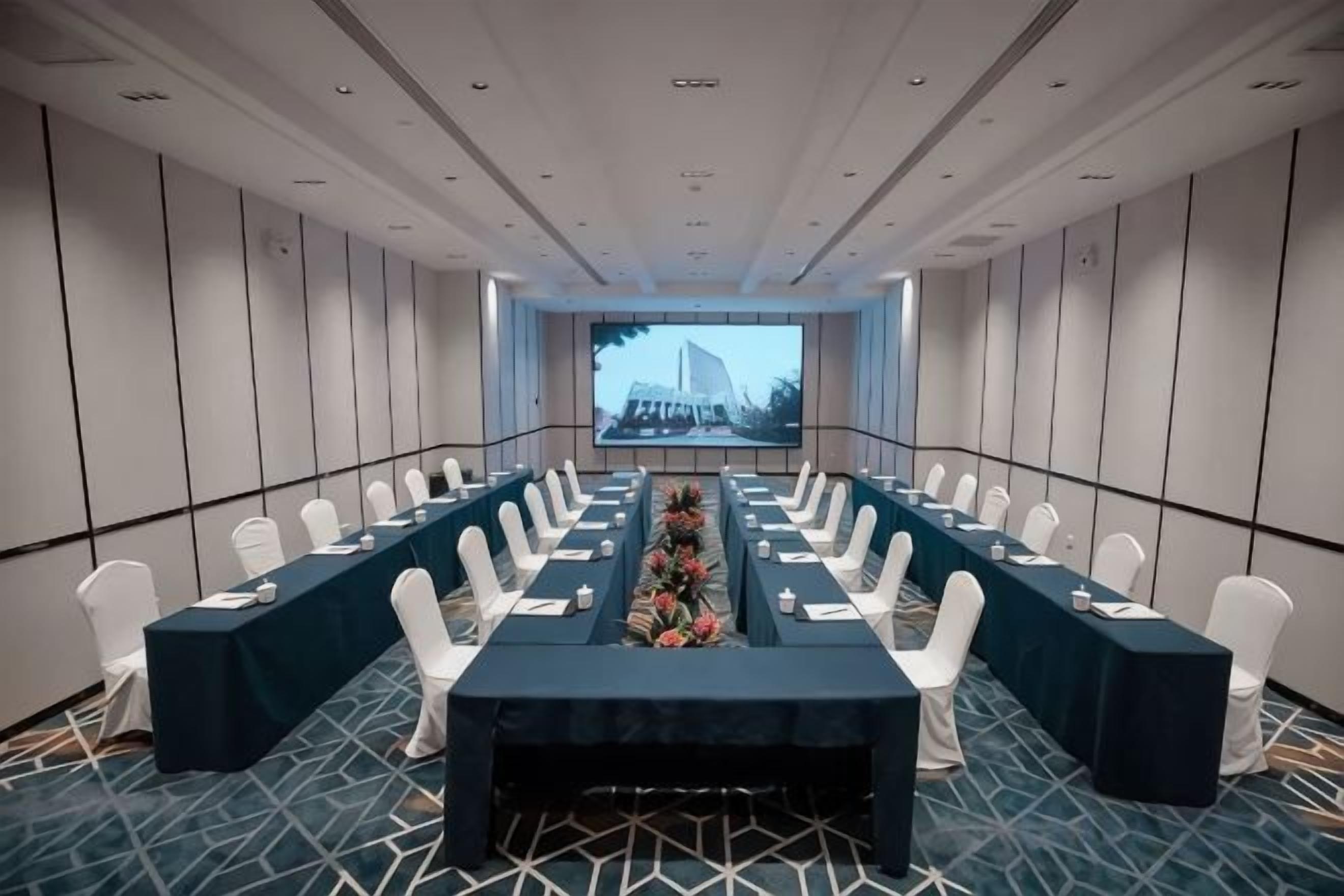 Un salón de conferencias con una larga mesa, sillas blancas y una pantalla al frente.