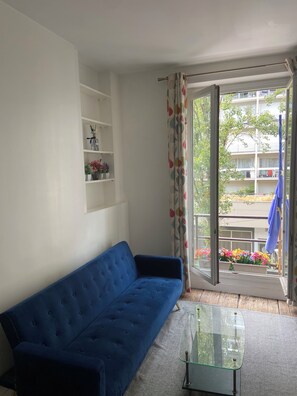 Ein Wohnzimmer mit einem blauen Sofa, einem Glastisch und Blick auf einen Balkon mit Blumen.