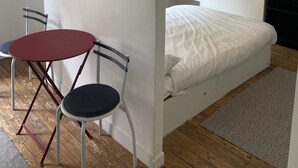 Ett litet rum med en säng, ett litet bord med stolar, en klocka på väggen och en hylla med växter.