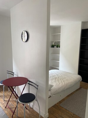 Ein gemütlicher Raum mit Bett, kleinem Tisch mit Stühlen, einer Wanduhr und einem Regal mit Pflanzen.