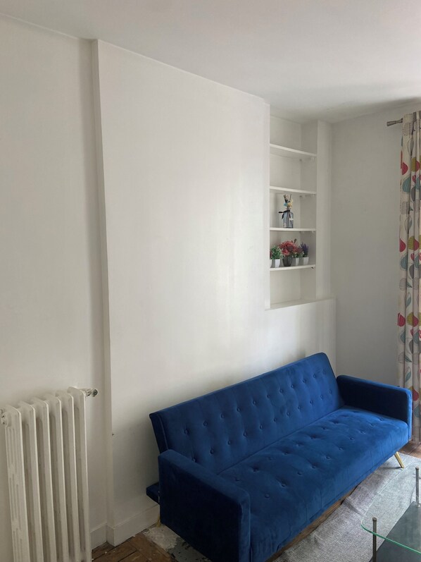 A living room with a blue sofa, white walls, a radiator, and a shelf with decorative items.
