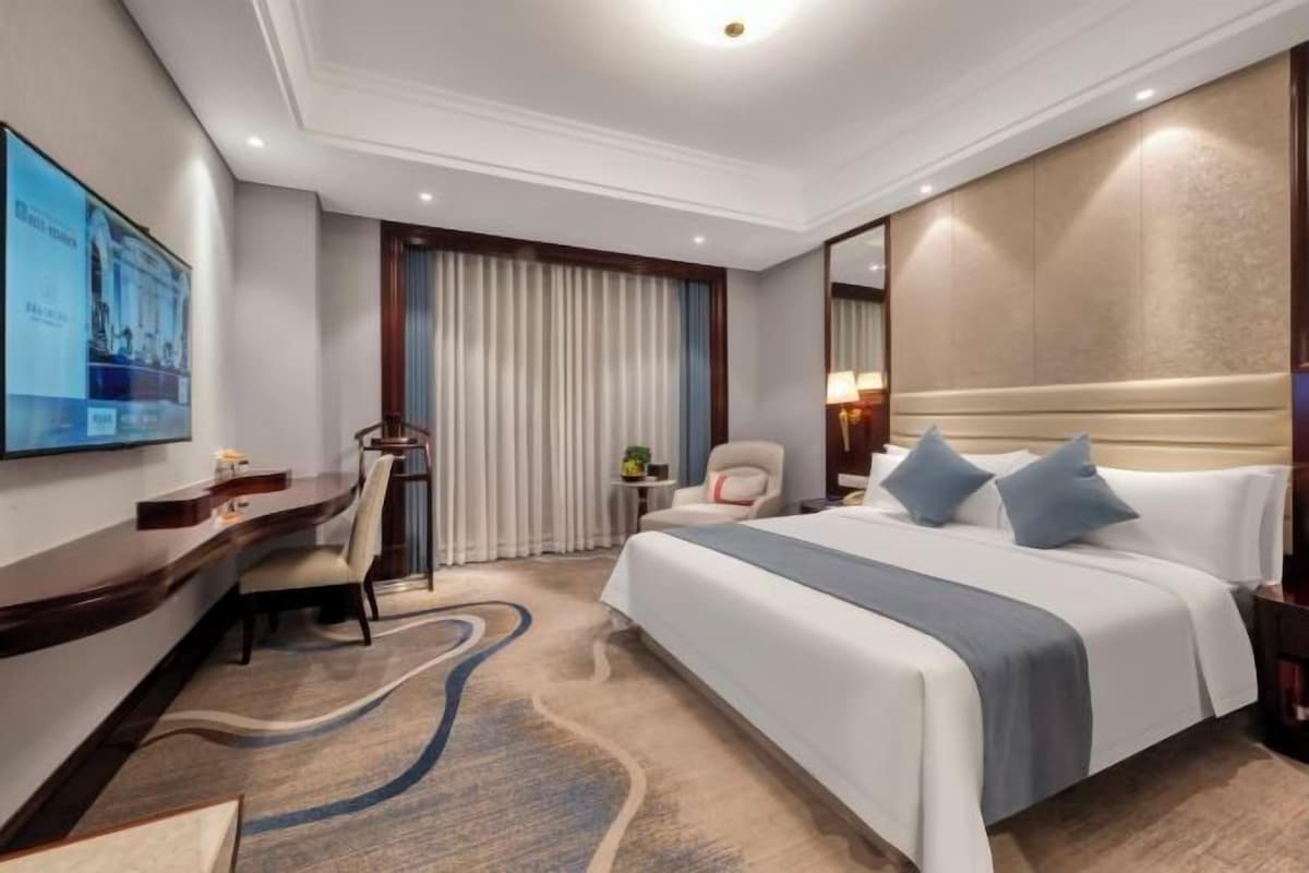 Habitación de hotel con cama, escritorio, silla, televisor y un ventanal con cortinas.