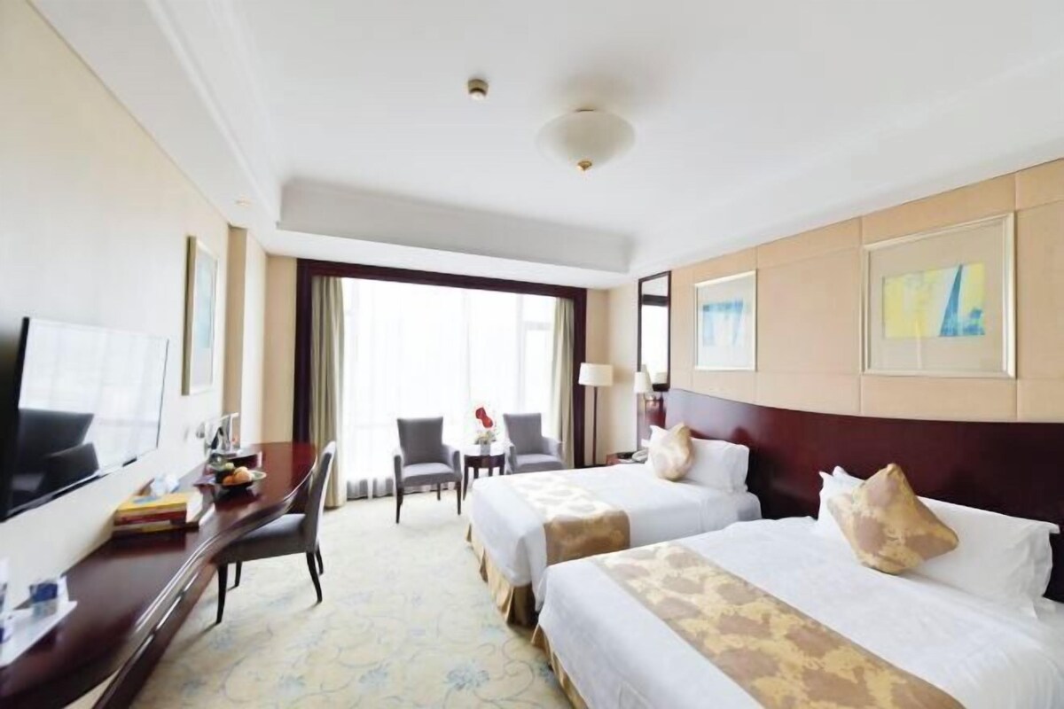A hotel room with two beds, a desk with a TV, and a large window.