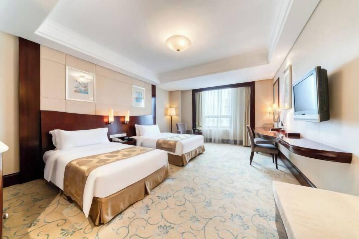 A hotel room with two beds, a desk, a TV, and a large window.