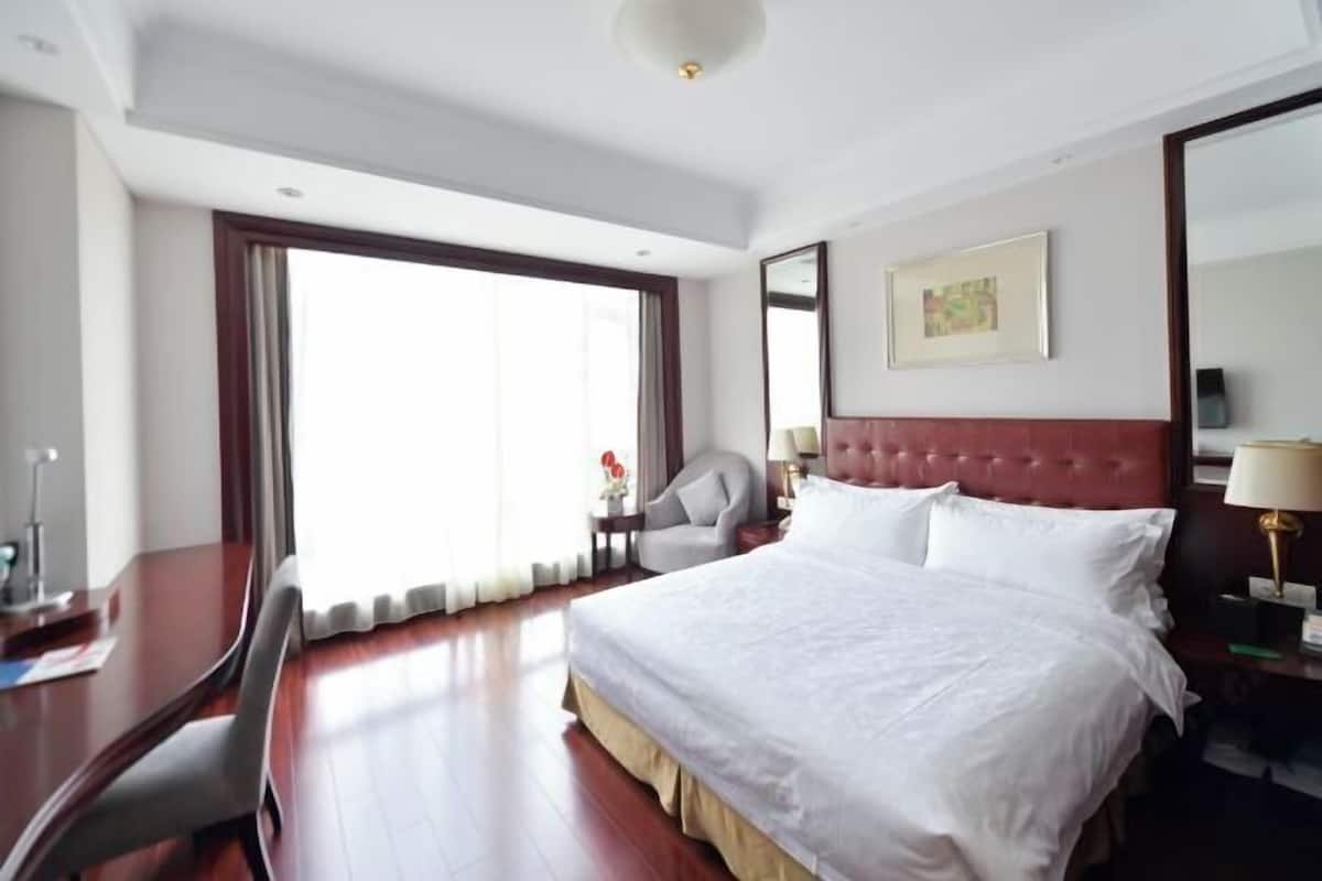 A hotel room with a large bed, a desk, a chair, a bedside table, and a window with curtains.
