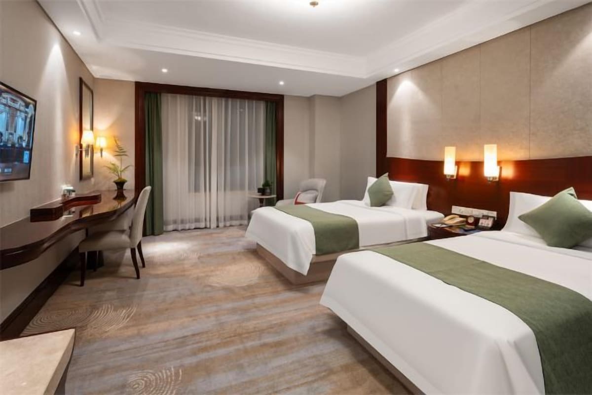 Habitación de hotel con dos camas, un escritorio, una silla, un televisor y un ventanal con cortinas.