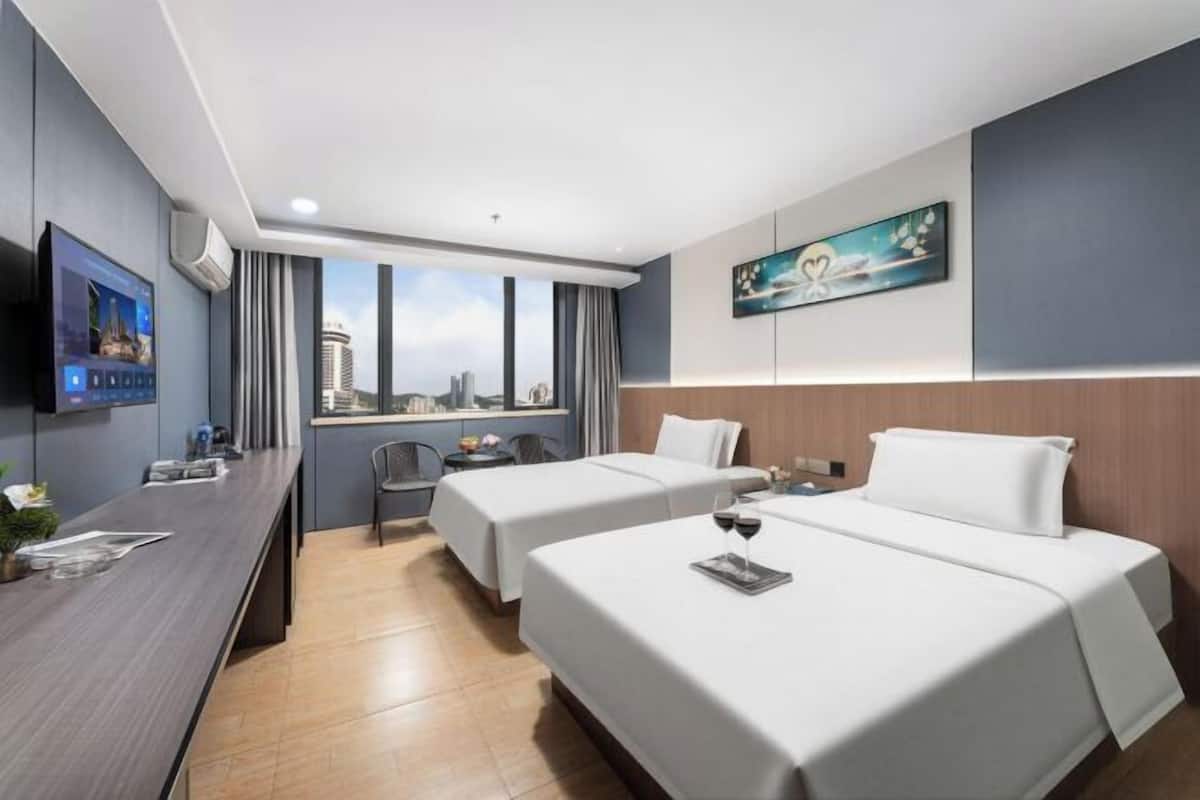 A hotel room with two beds, a desk with a flat-screen TV, a window with a city view, and a painting on the wall.