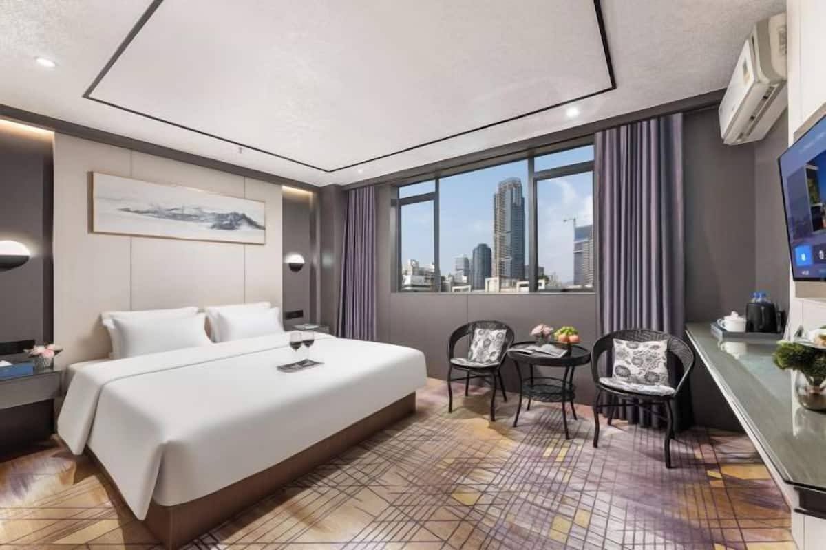 A hotel room with a large bed, a desk with a chair, a small table with a chair, and a city view through the window.