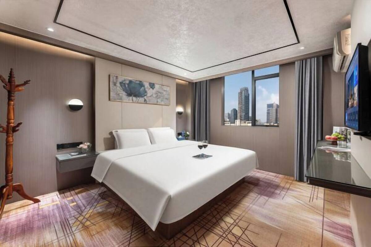 A modern hotel room with a large bed, a desk, a chair, a TV, and a city view.