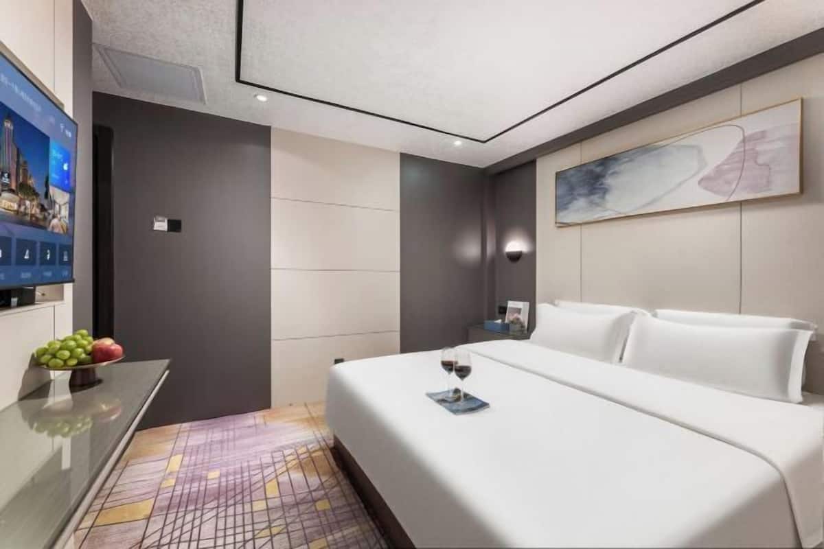 A modern hotel room with a bed, a bedside table with a fruit bowl, a wall-mounted TV, and a painting.