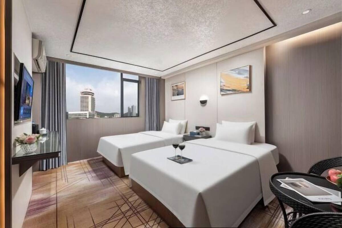 A hotel room with two beds, a desk with a chair, a small table with a chair, a TV, and a window with a city view.