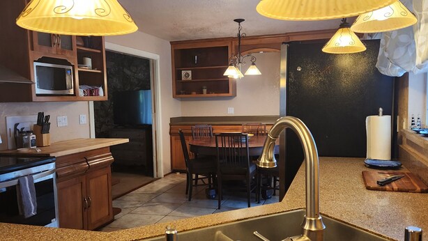 A kitchen with granite countertops, a stainless steel sink, and pendant lighting. There is a dining area with a table and chairs, and a microwave above the cabinets.