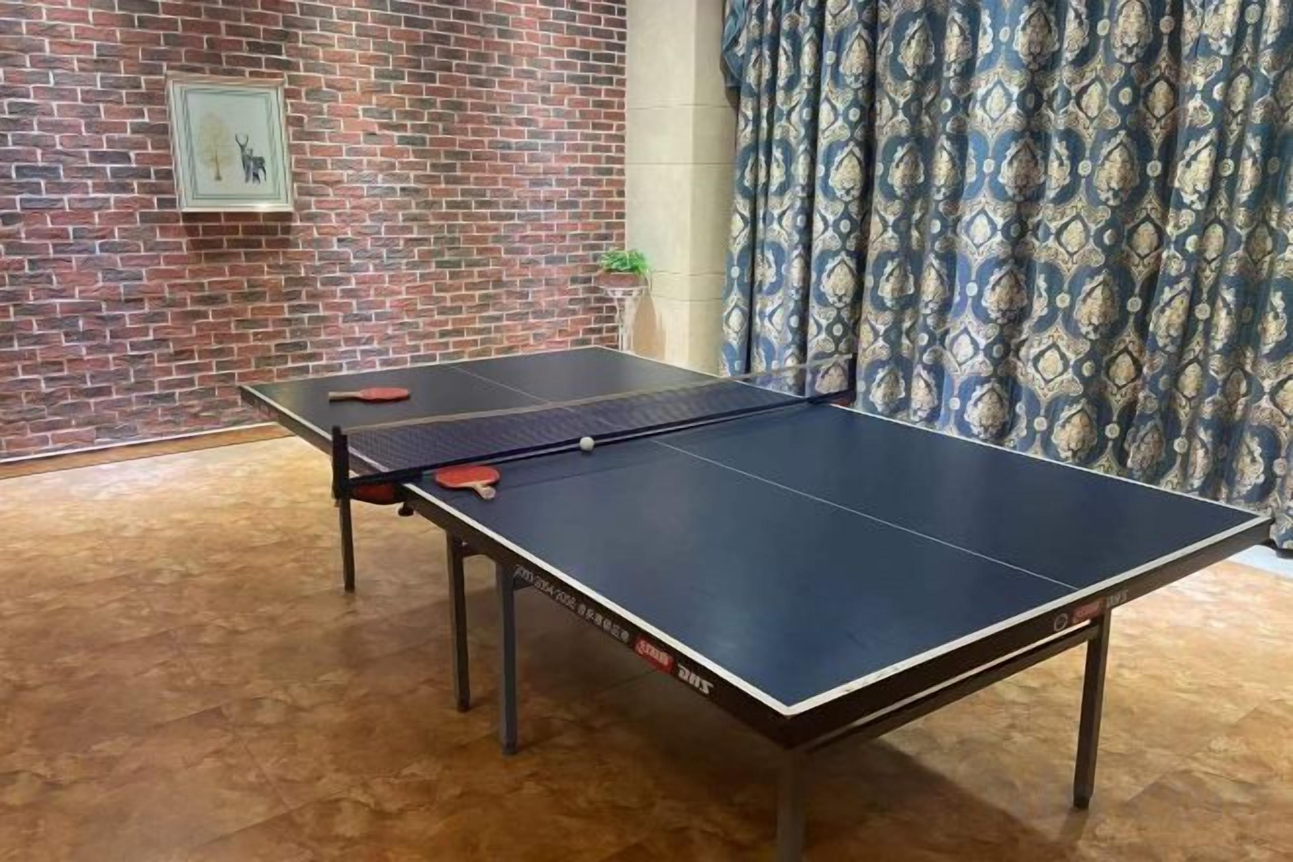 Una mesa de ping pong con paletas, una pared de ladrillo y una cortina estampada.