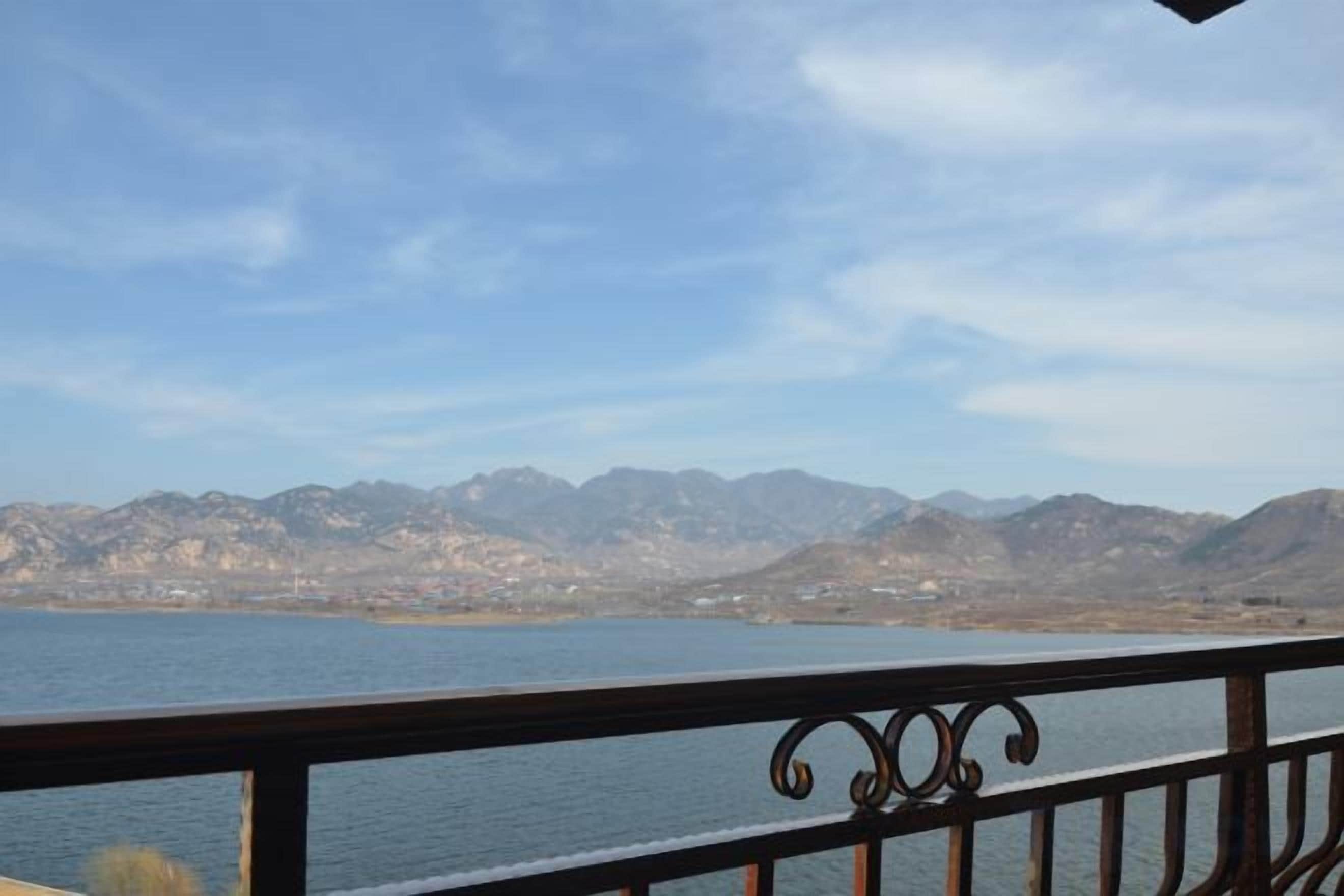 Vista de um lago com montanhas ao fundo, um corrimão de metal em primeiro plano e um céu claro.