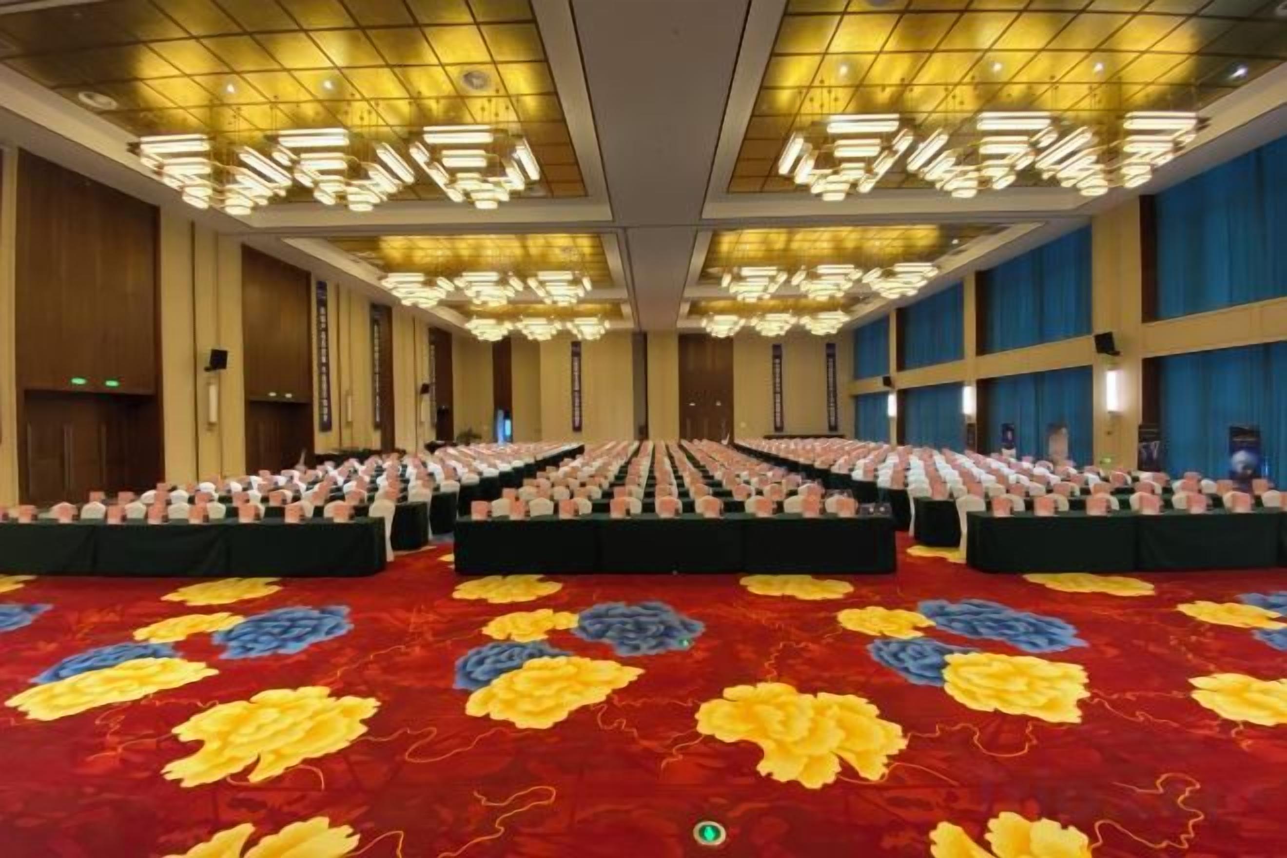 Un amplio salón de conferencias con filas de mesas y sillas, una alfombra estampada y un techo alto con luminarias.