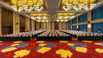 Un amplio salón de conferencias con filas de mesas y sillas, una alfombra estampada y un techo alto con luminarias.