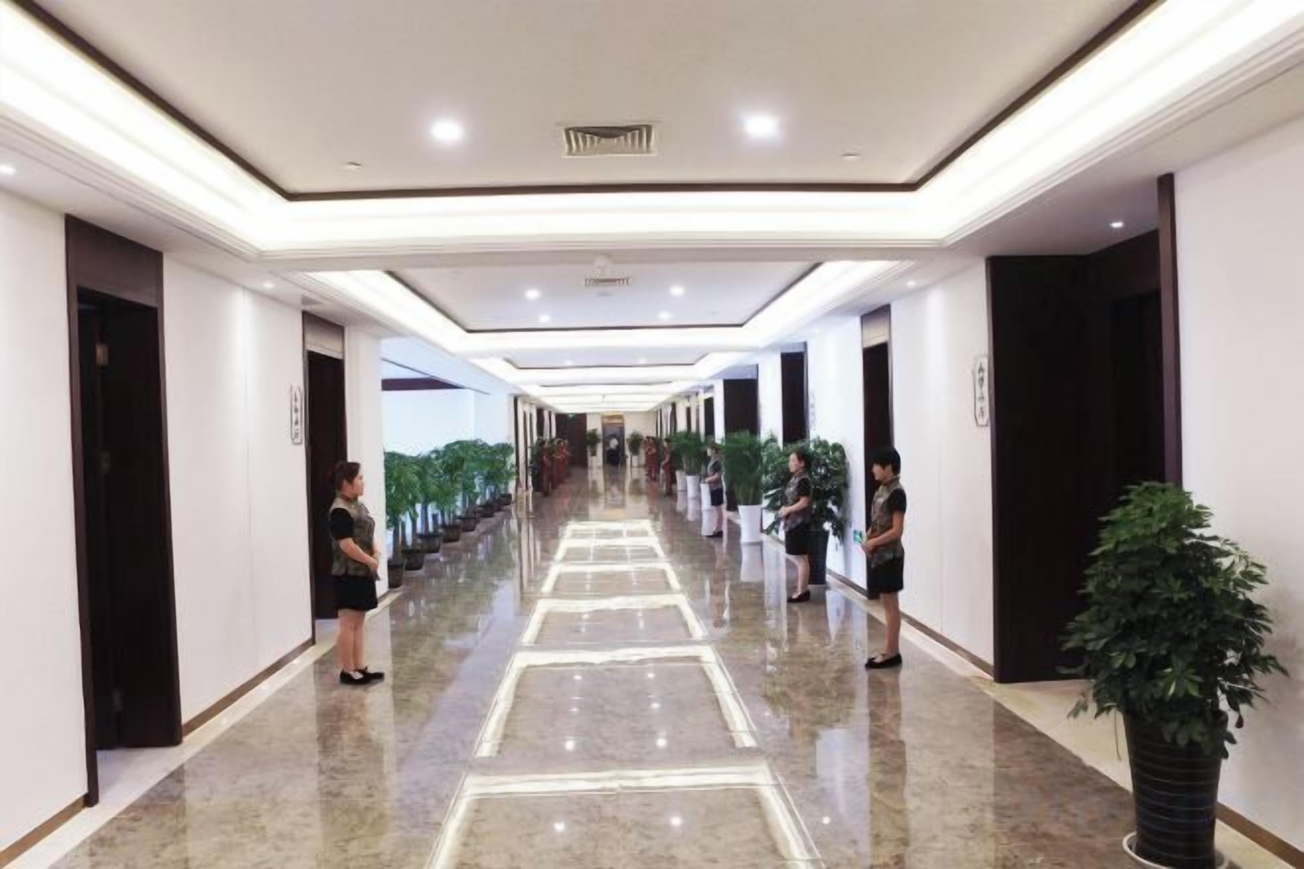 A hotel corridor with a polished floor, doors on both sides, and potted plants.