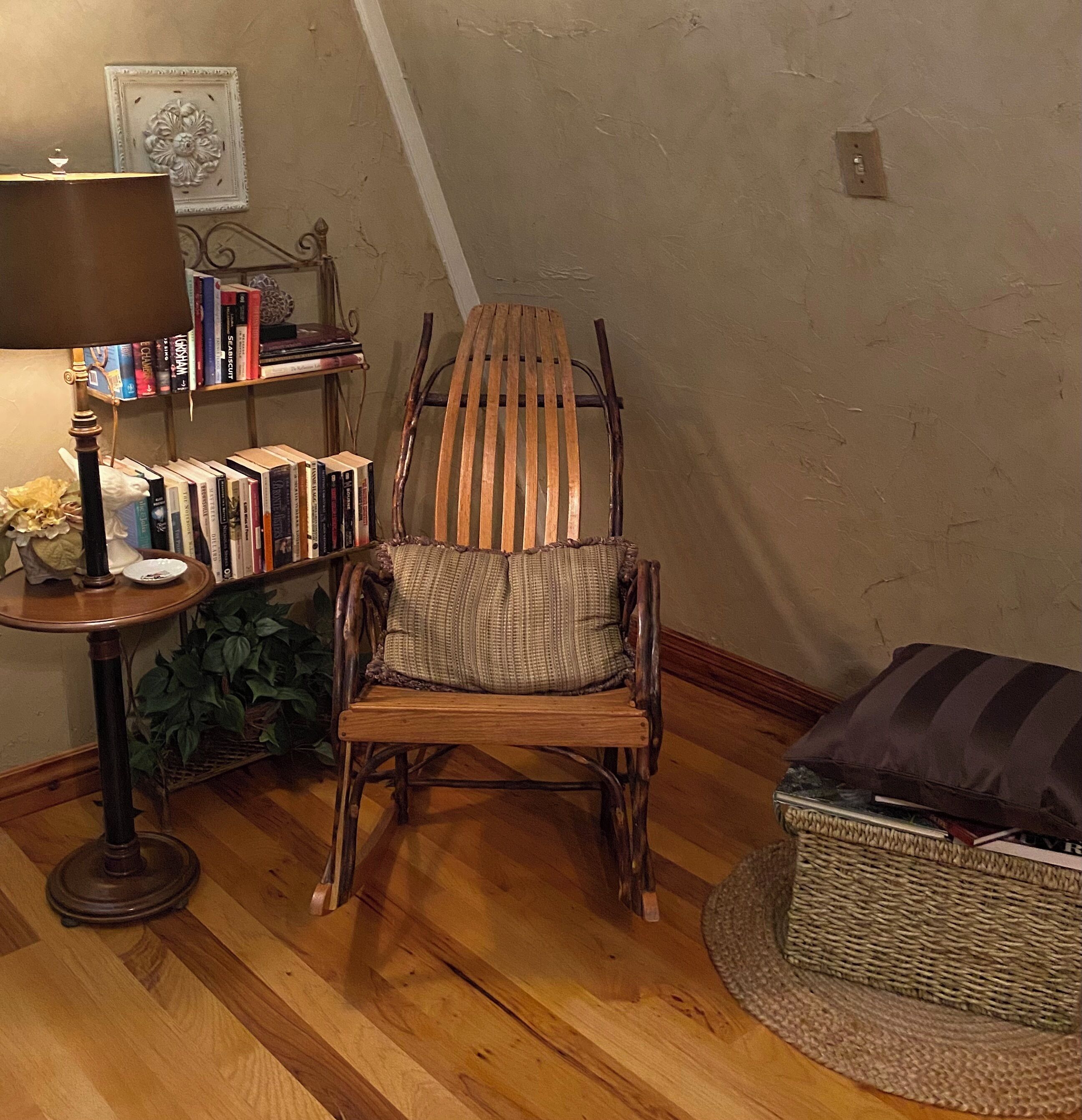 Una silla de madera con un cojín tejido, una mesita con una lámpara y una estantería llena de libros.