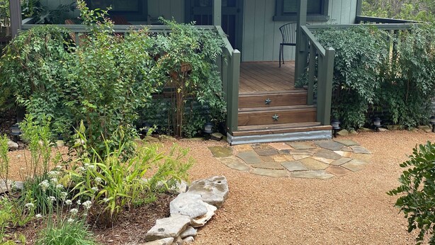 Une maison dotée d’un porche abrité, d’un chemin en pierre et d’un jardin où poussent différentes espèces de plantes.