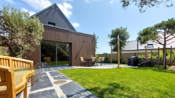 Ein modernes Haus mit einer großen Glasdoor, einer Terrasse mit einer Holzbank und einem gepflegten Rasen.