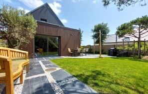 Une maison moderne dotée d’une grande porte vitrée, d’un patio avec un banc en bois et d’une pelouse bien entretenue.