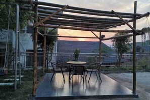 Un espace extérieur aménagé, abrité et doté de tables et de chaises, ainsi que d’une pergola, offrant une vue sur une maison dont le toit est recouvert de tuiles.