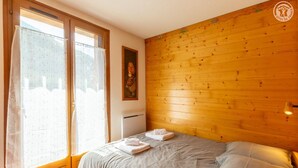 Quarto com cabeceira de madeira, cama com roupa de cama cinza, janela com cortinas e um pôster na parede.