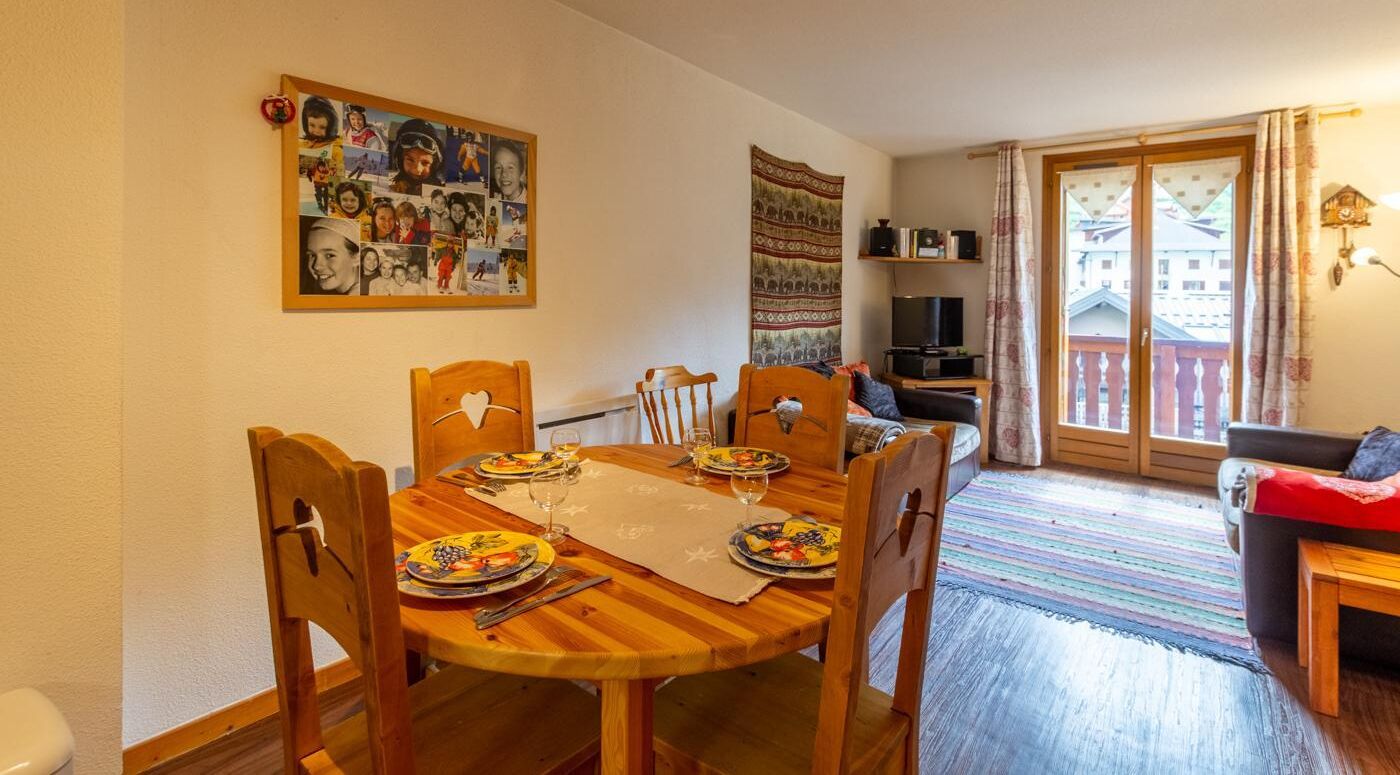 A dining area with a wooden table set for four, a wall with a collage of photos, and a view of a balcony with a railing.