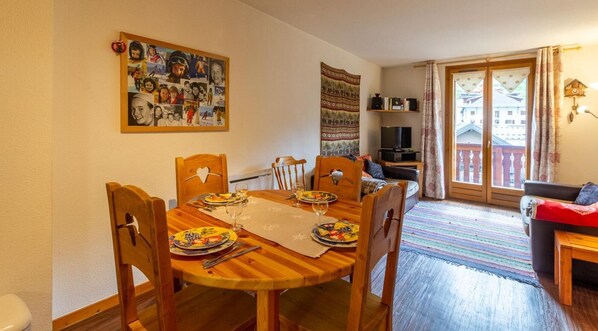A dining area with a wooden table set for four, a wall with a collage of photos, and a view of a balcony with a railing. - LE REPAIRE DES MARMOTTES (VALLOIRE)