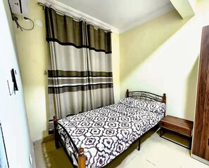 Ein Einzelbett mit einem gemusterten Bettüberwurf, eine Holzbank, ein Fenster mit Vorhängen und eine Tür mit Griff.
