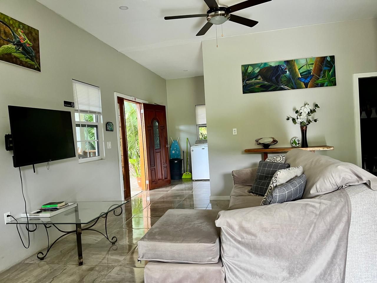 A living room with a flat-screen TV, a glass coffee table, a sofa with cushions, and artwork on the walls.