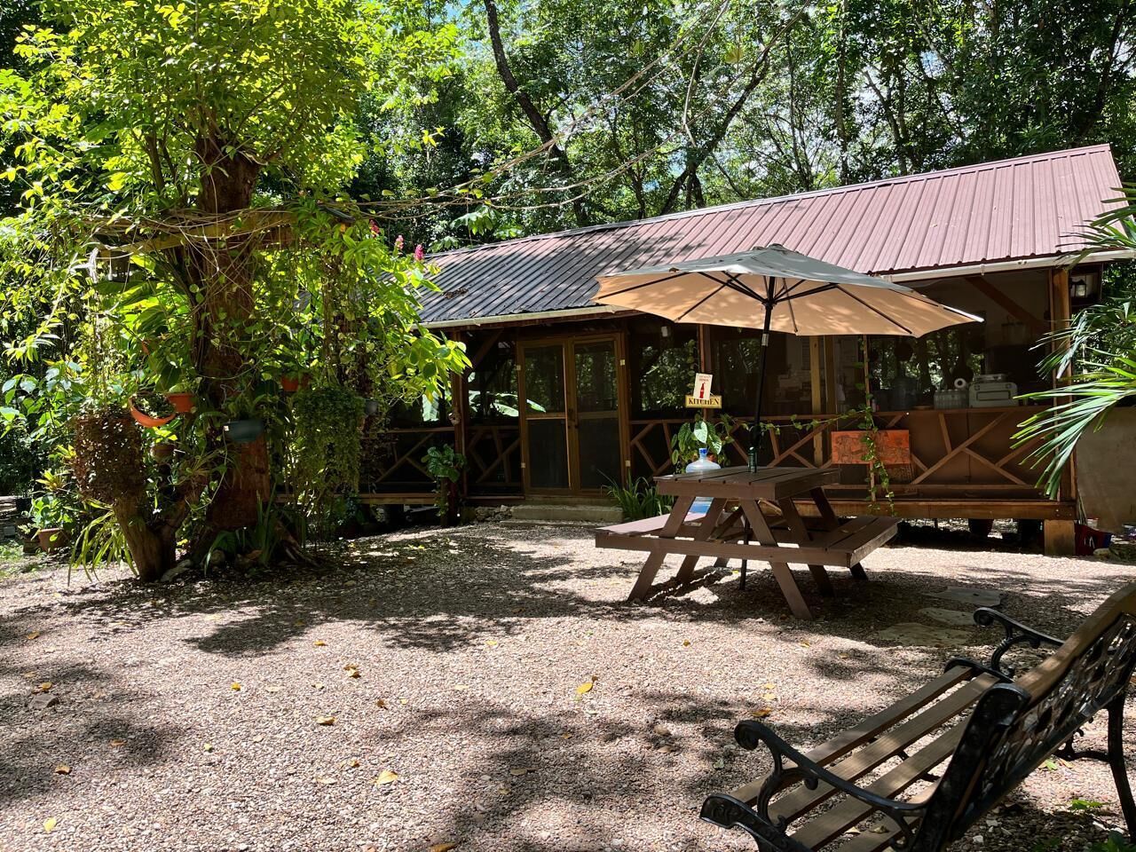 A covered outdoor seating area with a picnic table and benches, surrounded by trees and a gravel ground.