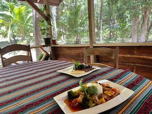 Une table en bois recouverte d’une nappe tissée aux couleurs vives, deux assiettes remplies de nourriture et, par la fenêtre, une vue sur une forêt.