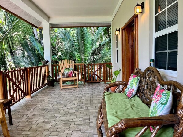 A covered porch with a wooden bench and a sofa, surrounded by a wooden railing and potted plants.