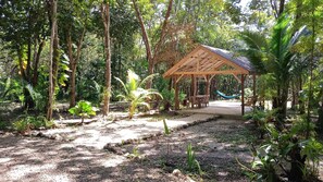 Un mirador de madera con techo de paja, rodeado de exuberante follaje tropical y una hamaca.