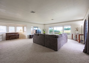 A spacious living room with a piano, a sectional sofa, and a large window offering a view of the outdoors. - Steps to beach! Pet friendly. (Crescent City)