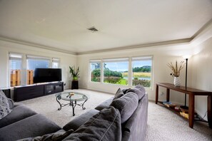 A living room with a large flat-screen TV, a sofa, a coffee table, and a view of a green landscape through large windows. - Steps to beach! Pet friendly. (Crescent City)