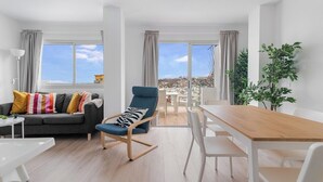 A modern living room with a sofa, a blue armchair, a wooden dining table, and a view of a cityscape through large windows.