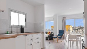 A modern kitchen with white cabinets and a wooden countertop, a dining area with a table and chairs, and a view of buildings through large windows.