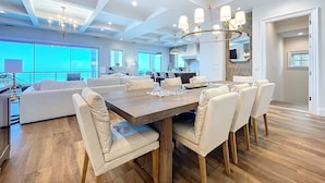 A spacious living room with a large wooden dining table, white upholstered chairs, and a chandelier.