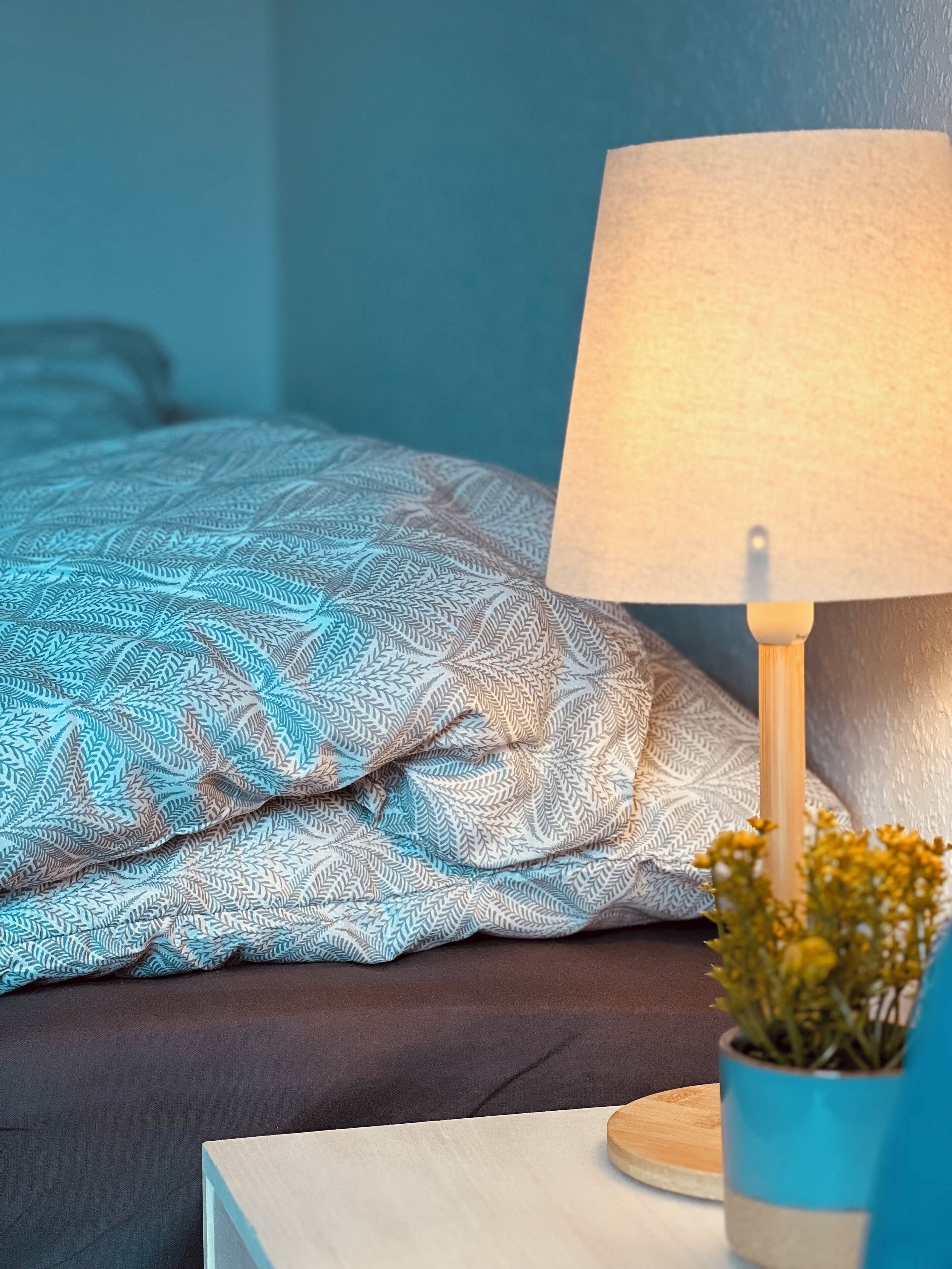 Un lit bien fait, avec une literie à motifs, une table de chevet avec une lampe et une petite plante en pot.