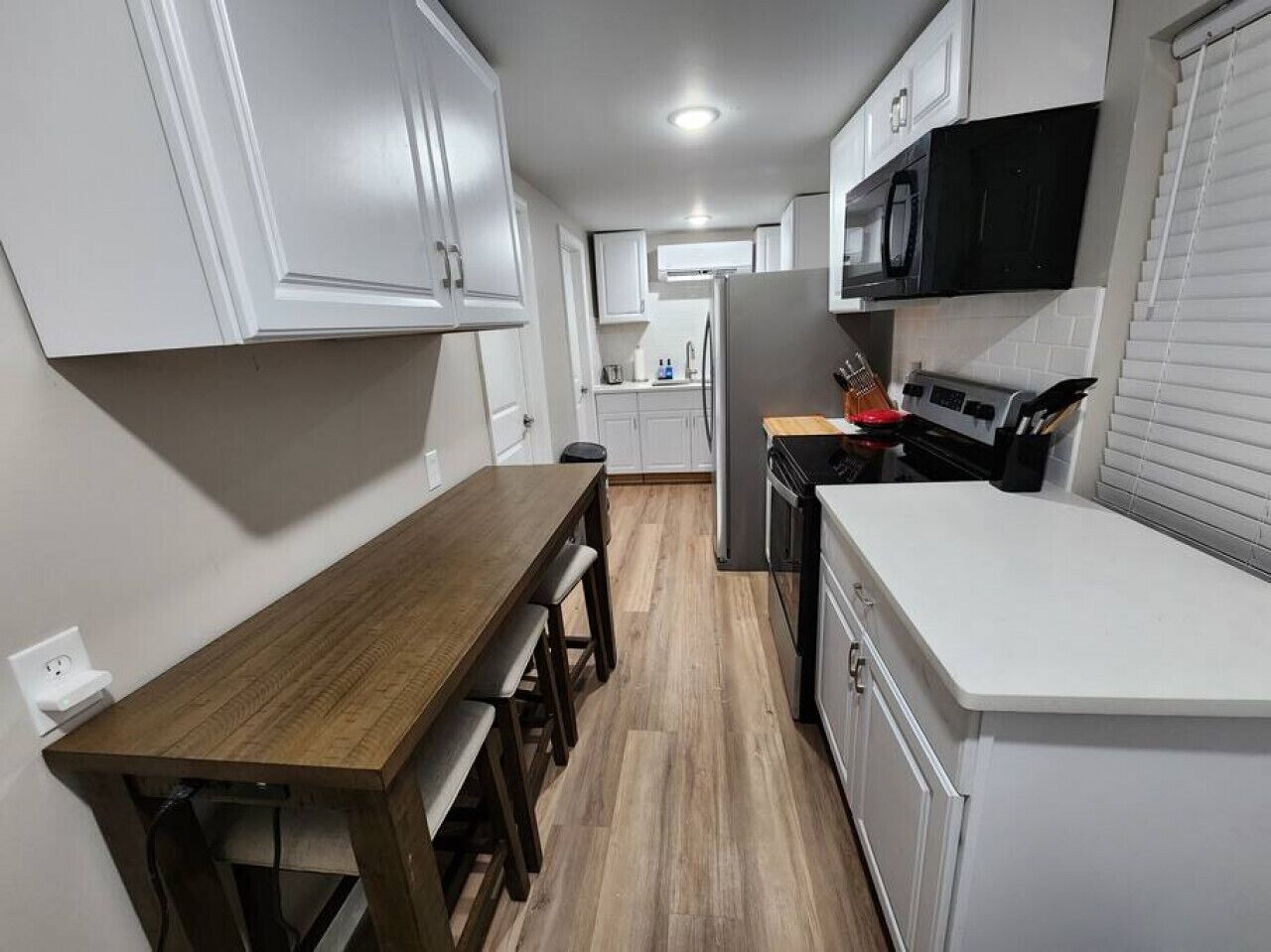 Een kleine keuken met witte kastjes, een houten eettafel en een magnetron.