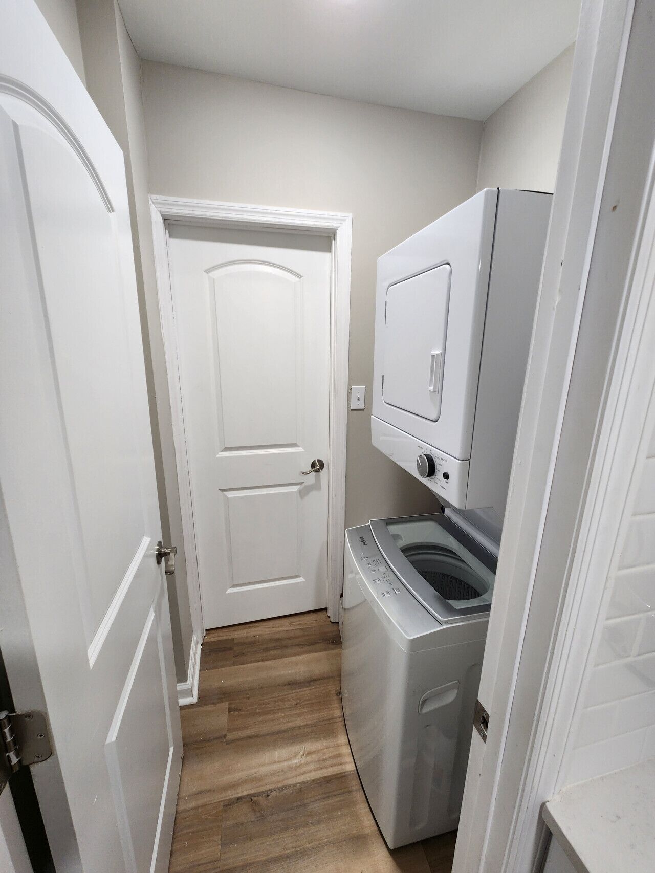 Pequeña lavandería con lavadora y secadora, una puerta blanca y piso de madera.