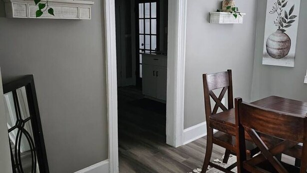 A dining area with a dark wood floor, a white rug, and a wooden dining table with chairs. There are two white shelves with plants and decorative items. - The White House 2 BR 1 BA (Savannah)