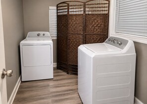A laundry room with two white front-loading washers and dryers, a brown woven partition, and a window with blinds.