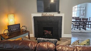 A living room with a fireplace, a TV mounted above it, and a dining area visible through an open doorway. - The White House 2 BR 1 BA (Savannah)