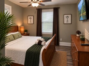 A bedroom with a bed, a nightstand, a dresser, a television, and a window with curtains.