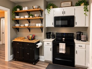 A kitchen with a black stove, white cabinets, and a wooden shelf with bowls and cups.