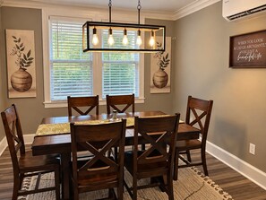 A dining room with a rectangular table, six wooden chairs, a hanging light fixture, and two windows with blinds.