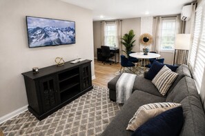 A living room with a grey sofa, a black entertainment unit, a flat-screen TV, a round dining table with chairs, and a potted plant.